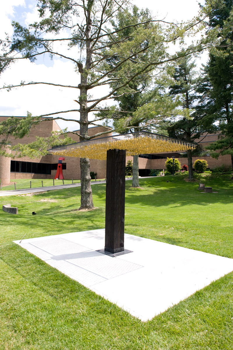 An outdoor sculpture featuring a thick, dark wooden pillar supporting a wide, flat, square canopy. The underside of the canopy is entirely densely covered with hundreds of small, hanging yellow objects. The structure stands on a white rectangular concrete pad in the middle of a manicured green lawn, surrounded by tall pine trees and campus buildings in the background.