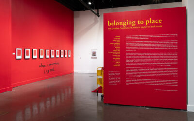 Gallery with red walls displaying artwork and text for "belonging to place" exhibition