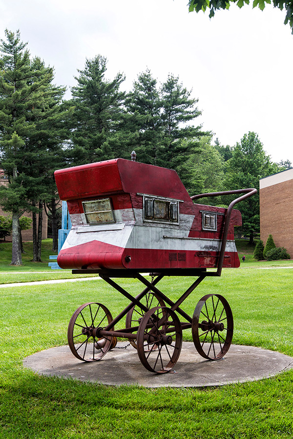 A whimsical, rustic outdoor sculpture of a vintage-style, high-wheeled baby carriage or stroller on a circular concrete base. The body of the carriage resembles a small camper or trailer painted with weathered red and white blocks, complete with tiny rectangular windows. The sculpture is set against a backdrop of a green lawn and dense pine trees under an overcast sky.