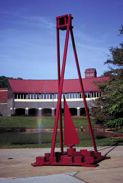 A tall, geometric red metal sculpture standing on a concrete plaza. The piece consists of a complex base made of blocky forms, a central sail-like triangle, and two long, slender beams that extend upward to meet at a small enclosed rectangular box high in the air. In the background, a small pond with a water fountain is visible in front of a large brick building with a red metal roof.