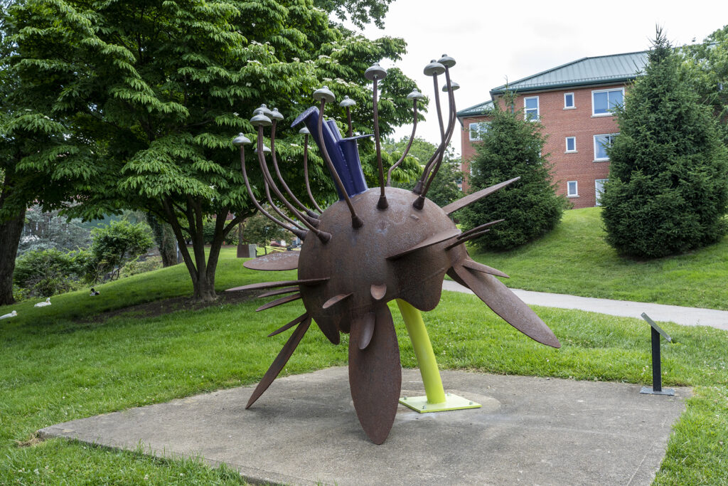 A large outdoor metal sculpture shaped like a textured, rusty sphere with long spikes, fin-like blades, and curved stalks topped with silver, cap-like elements, supported by a bright green post on a concrete pad.