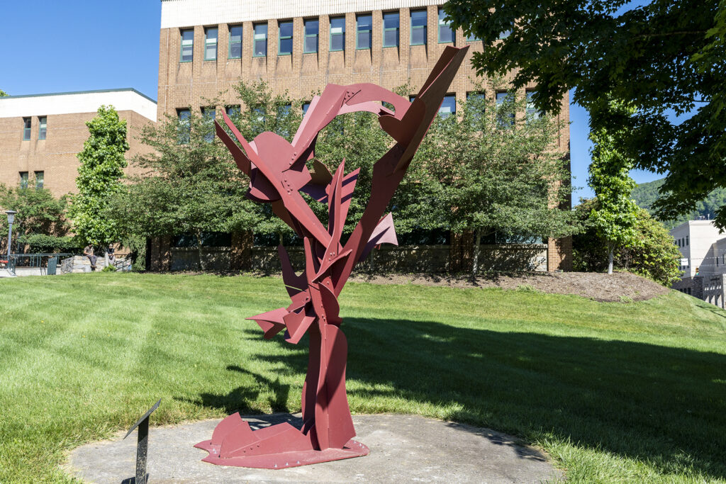 Abstract reddish-brown metal sculpture with sharp, wing-like geometric panels extending outward, installed on a circular concrete pad on a grassy lawn in front of a large leafy tree.