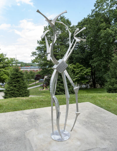 Tall, abstract silver metal sculpture featuring slender, angular bars and curved elements that twist upward from a circular base, meeting at a central diamond-shaped metal plate before branching toward the sky.