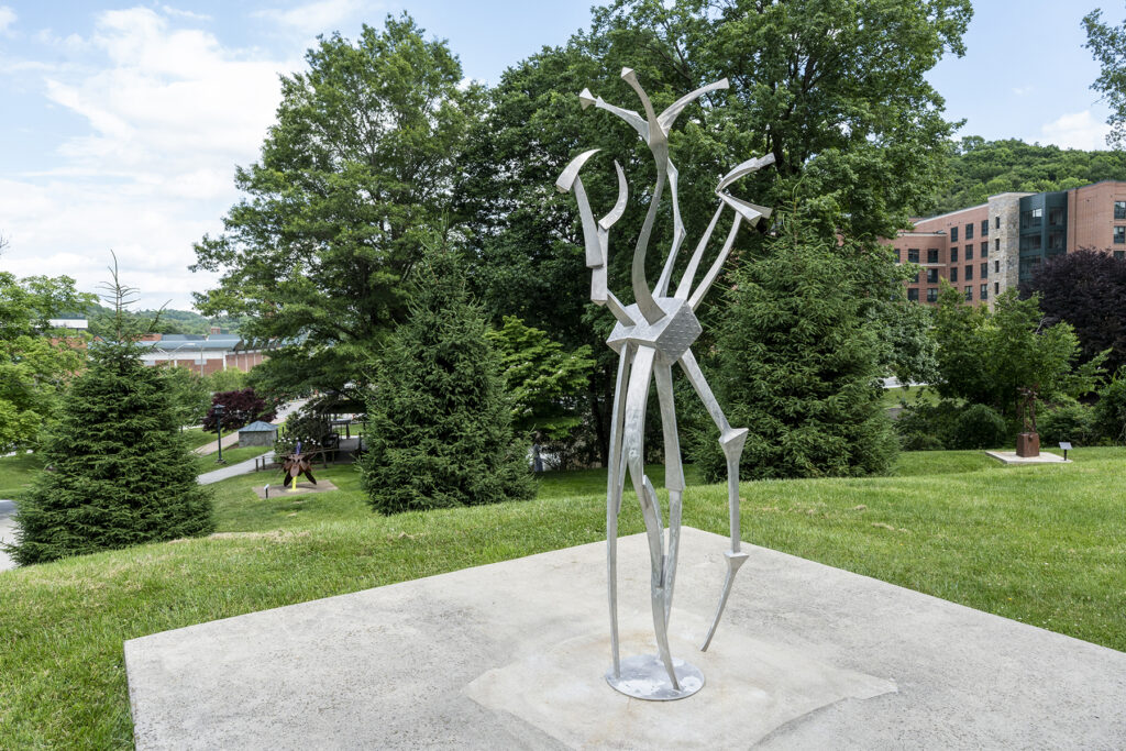 Tall, abstract silver metal sculpture featuring slender, angular bars and curved elements that twist upward from a circular base, meeting at a central diamond-shaped metal plate before branching toward the sky.