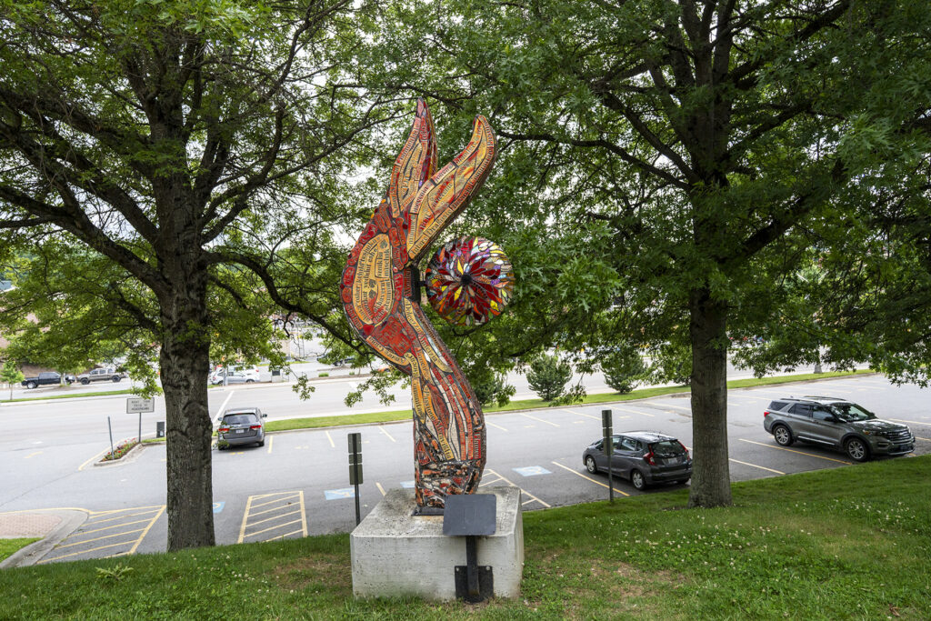 A vertical, flame-shaped mosaic sculpture covered in red, orange, and gold tiles with embedded text, featuring a central multicolored stained-glass sphere, positioned on a concrete pedestal between two large trees overlooking a parking lot.