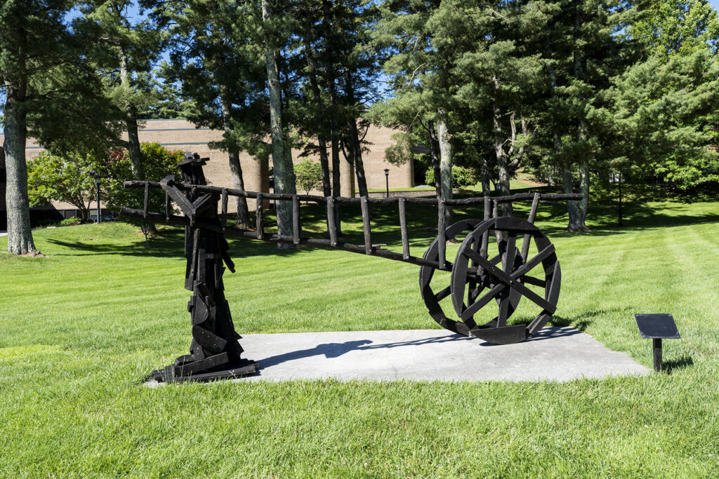 A black wooden sculpture depicting a stylized human figure pulling a long, horizontal ladder-like cart with a single large, spoked wheel, installed on a concrete pad on a sunny campus lawn.