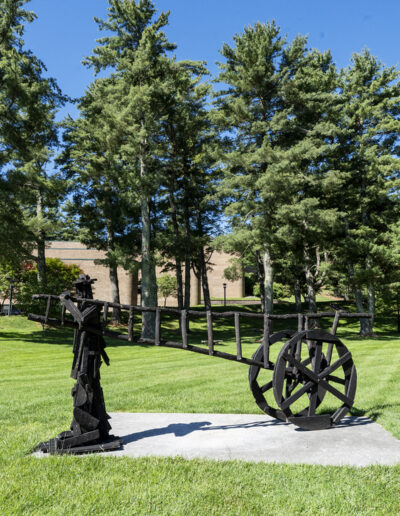 A black wooden sculpture depicting a stylized human figure pulling a long, horizontal ladder-like cart with a single large, spoked wheel, installed on a concrete pad on a sunny campus lawn.