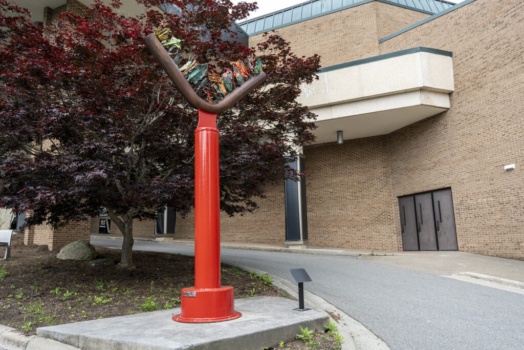 An outdoor sculpture featuring a thick, bright red metal pillar supporting a dark, crescent-shaped trough filled with colorful, abstract glass elements, situated on a concrete base next to a dark red maple tree.