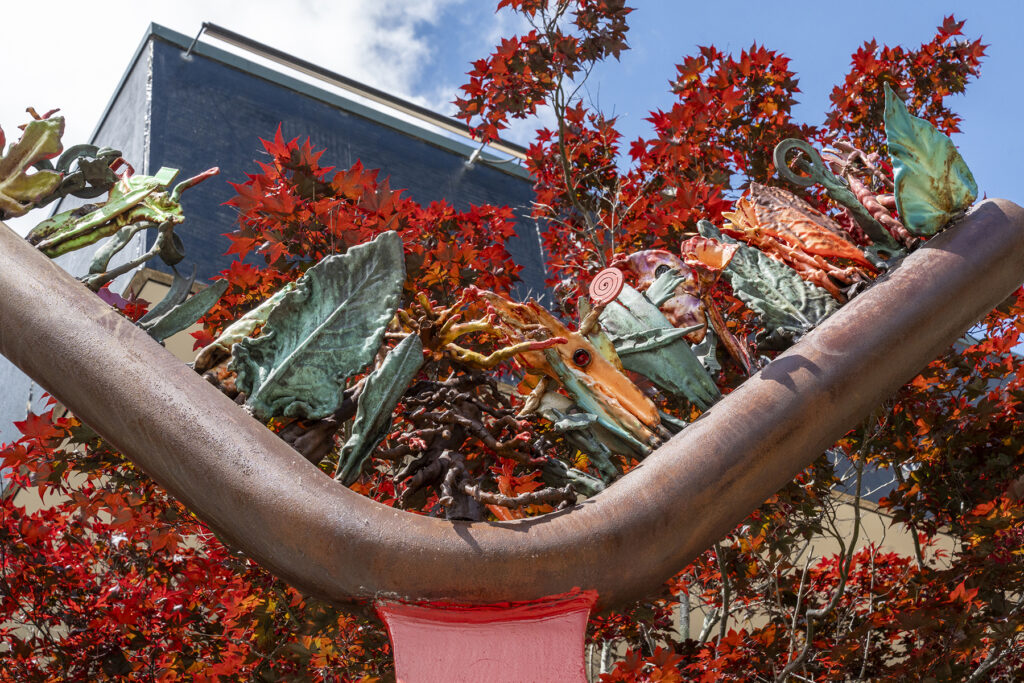 An outdoor sculpture featuring a thick, bright red metal pillar supporting a dark, crescent-shaped trough filled with colorful, abstract glass elements, situated on a concrete base next to a dark red maple tree.