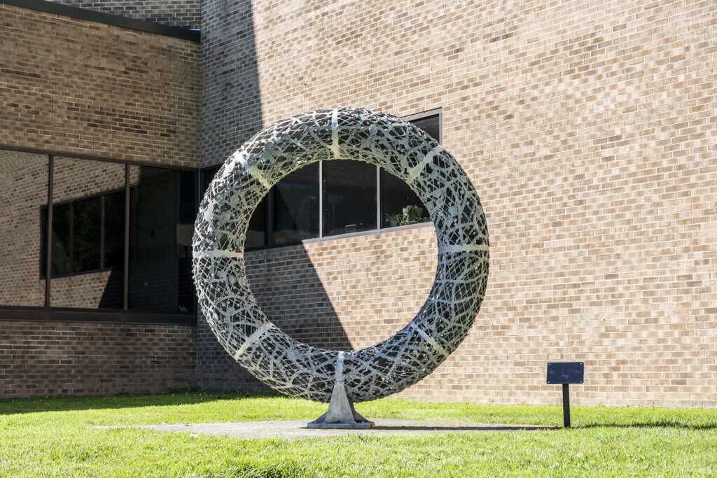 A large, circular metal ring sculpture with an intricate, open latticework or woven pattern, anchored to a small base on a grassy area in front of a brick building.
