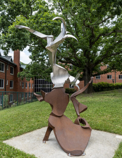 A large, circular metal ring sculpture with an intricate, open latticework or woven pattern, anchored to a small base on a grassy area in front of a brick building.