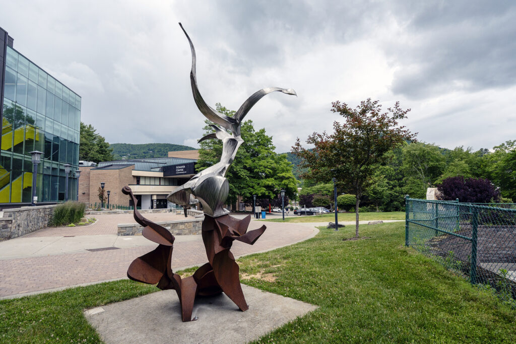 A large, circular metal ring sculpture with an intricate, open latticework or woven pattern, anchored to a small base on a grassy area in front of a brick building.