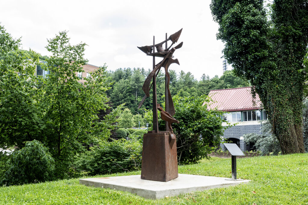 A tall, rusted iron sculpture featuring abstract, blade-like geometric shapes and sweeping metal ribbons branching upward from a solid rectangular pedestal, set on a concrete pad surrounded by lush green trees.
