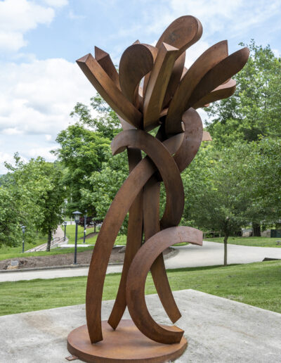 A large, rusted steel sculpture resembling a stylized flower, featuring thick, curved metal bands that twist upward from a circular base to form an open cluster of petal-like shapes at the top.