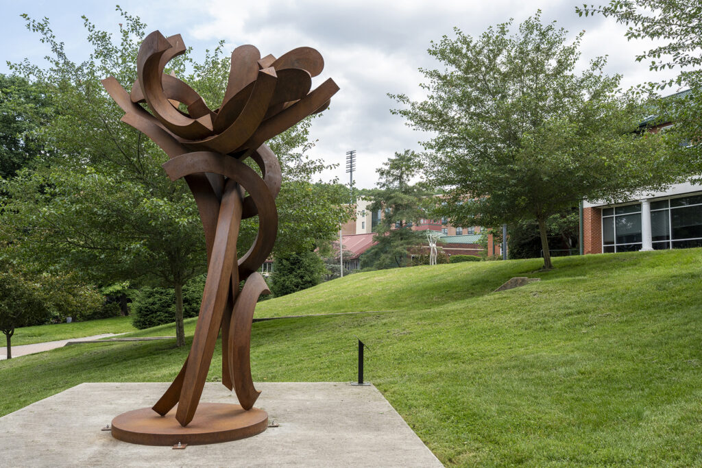 A large, rusted steel sculpture resembling a stylized flower, featuring thick, curved metal bands that twist upward from a circular base to form an open cluster of petal-like shapes at the top.